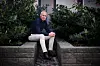 Person sitter på en steinkant foran busker og blomstrende planter i et utendørs hageområde.